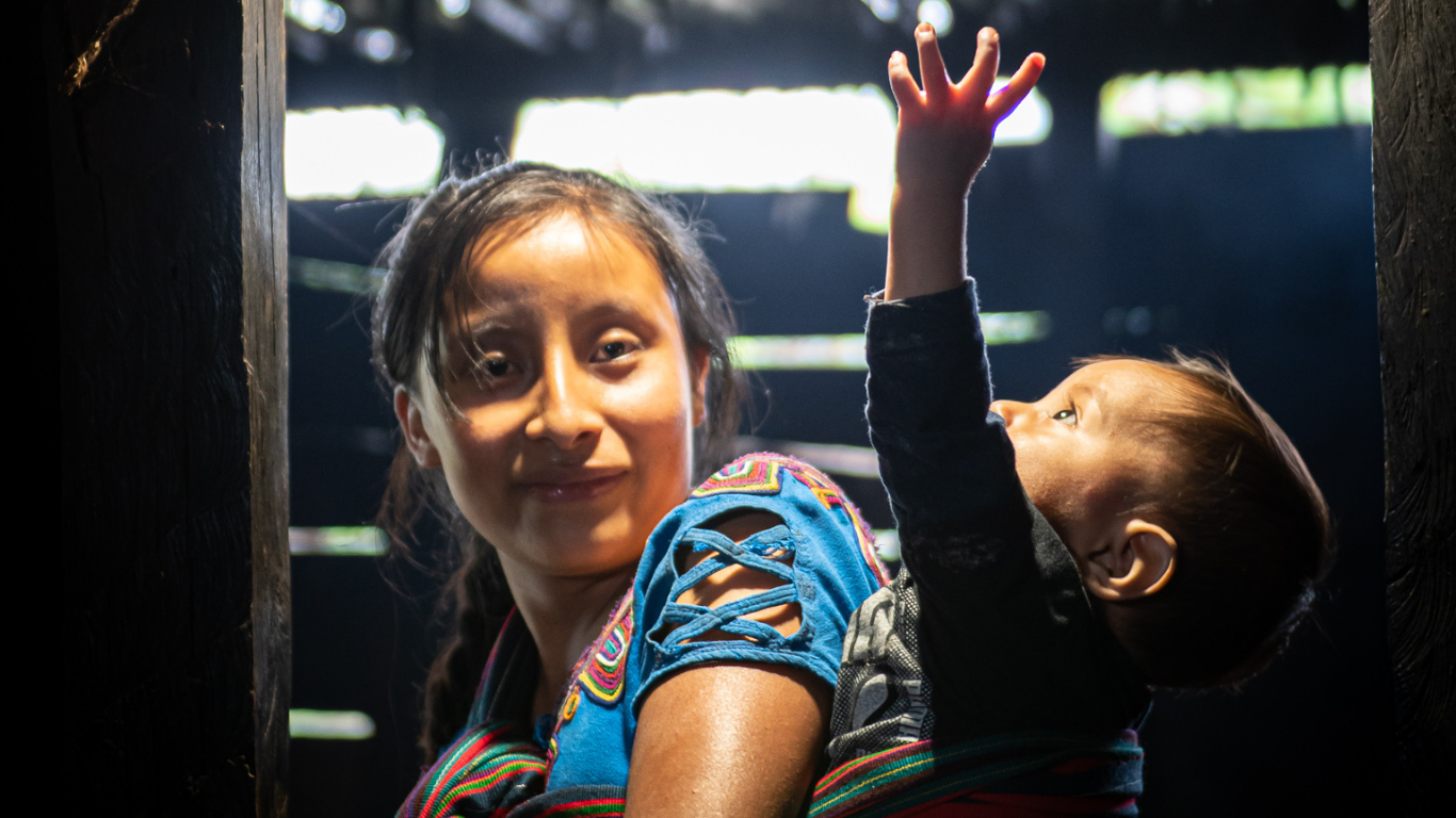 A mother carries her son on her back. He is reaching up to the ceiling towards a new solar light that has been installed. Empowering rural women is essential to increase access to energy globally. She is looking proud and excited.
